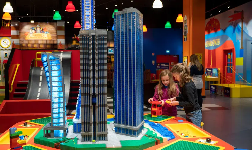 Two young girls are engaged in creative play, constructing miniature buildings at Legoland Discovery Centre. A vibrant, interactive LEGO city display with skyscrapers surrounds them, offering a hands-on experience. Shopping discounts for disabled people, family-friendly attractions, and accessible entertainment.