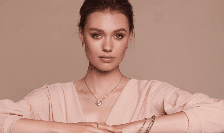 A woman with light skin, wearing a light pink blouse, poses against a plain background. She has brown hair and wears a necklace, earrings, and bracelets.