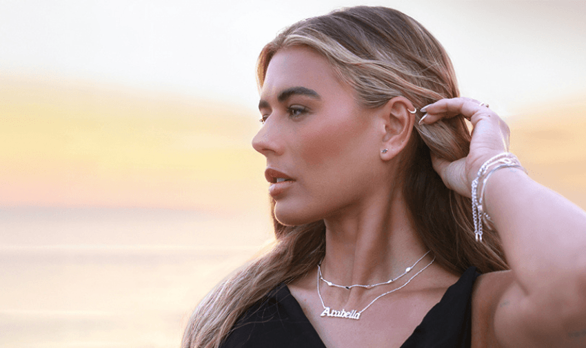 A woman wearing a silver name necklace and delicate bracelets, gazing into the distance with a golden sunset in the background. Showcasing Purpl's disabled discounts for elegant and personalised jewellery from Say It With in the UK.