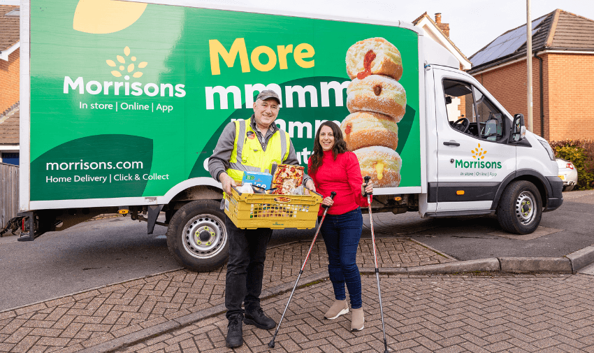 A Morrisons delivery driver hands over groceries to a woman in a red coat, with a Morrisons delivery van parked outside a suburban home. Disabled grocery delivery offers UK.