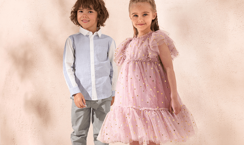 Two children are standing side by side against a light-coloured background. The child on the left is wearing a white dress shirt and grey pants, while the child on the right is in a pink, frilly dress with gold polka dots. Both children are smiling.