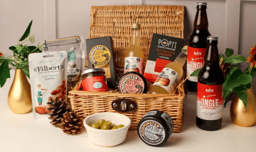 A wicker basket filled with assorted gourmet items, including cheese, chutney, ale bottles, almonds, crackers, and a jar of olives. It's surrounded by decorative pine cones and small vases with plants.