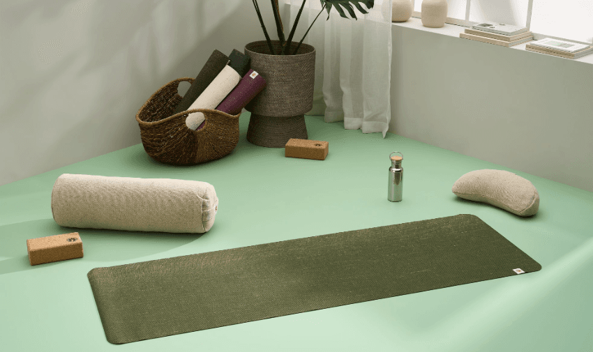 A peaceful yoga setup featuring a green eco-friendly yoga mat, natural cotton bolster, meditation cushion, and cork yoga blocks in a softly lit space. A woven basket holds additional rolled-up mats, adding to the minimalist and sustainable aesthetic. Ideal for relaxation, meditation, and wellness. Includes SEO for accessible fitness discounts and mobility-friendly yoga accessories.