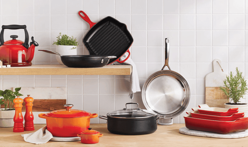 A kitchen scene with various Le Creuset cookware items: a red tea kettle, two red bell pepper-shaped grinders, an orange Dutch oven, a small orange pot, a black grill pan, a black frying pan, a stainless steel pan, red baking dishes, utensils, and potted herbs.