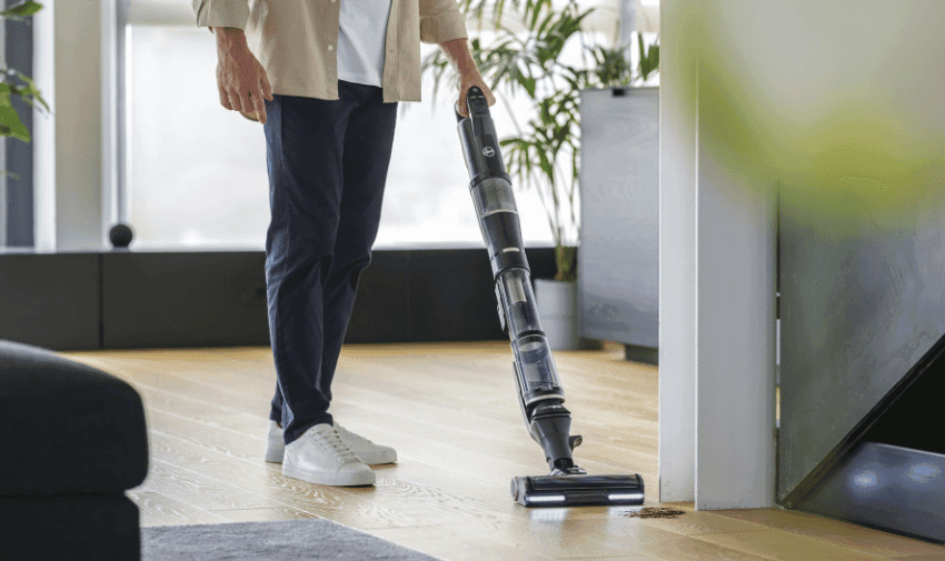 A person wearing white sneakers, blue pants, and a beige shirt is vacuuming a light wooden floor with a cordless vacuum cleaner. The room appears modern and bright, with sunlight streaming in. Some furniture and a potted plant are partially visible in the background.