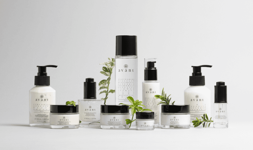 A collection of Avant skincare products displayed against a clean white background. The range includes bottles and jars of various sizes, featuring sleek black and white packaging with the Avant logo. Fresh green leaves are placed around the products, enhancing the natural and luxurious feel of the display