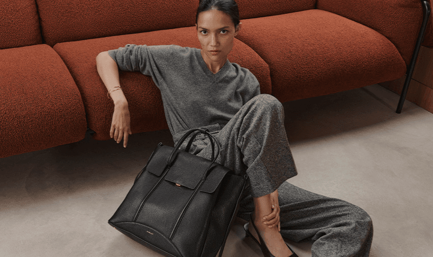 A person in a grey outfit sits on the floor, leaning against an orange sofa. They have a neutral expression and are holding a large black handbag placed beside them. The setting appears modern and minimalist