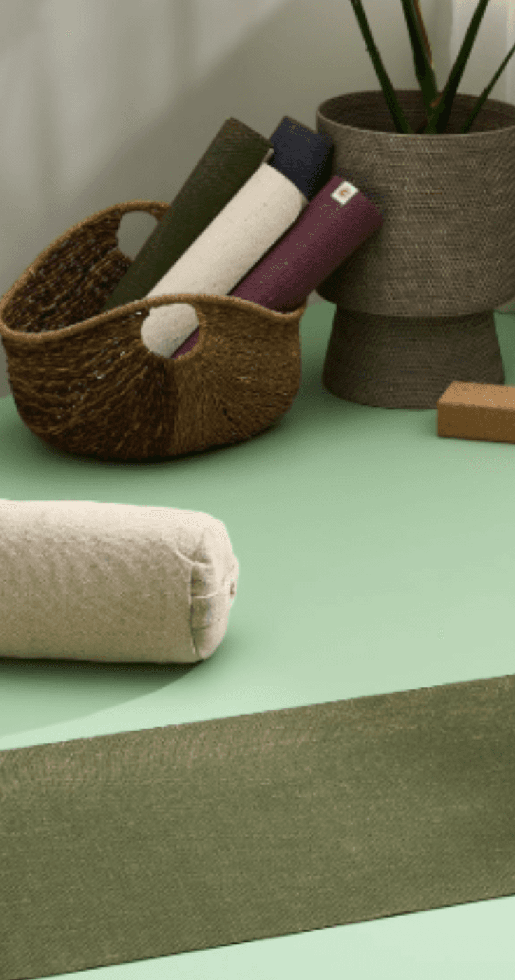 A peaceful yoga setup featuring a green eco-friendly yoga mat, natural cotton bolster, meditation cushion, and cork yoga blocks in a softly lit space. A woven basket holds additional rolled-up mats, adding to the minimalist and sustainable aesthetic. Ideal for relaxation, meditation, and wellness. Includes SEO for accessible fitness discounts and mobility-friendly yoga accessories.