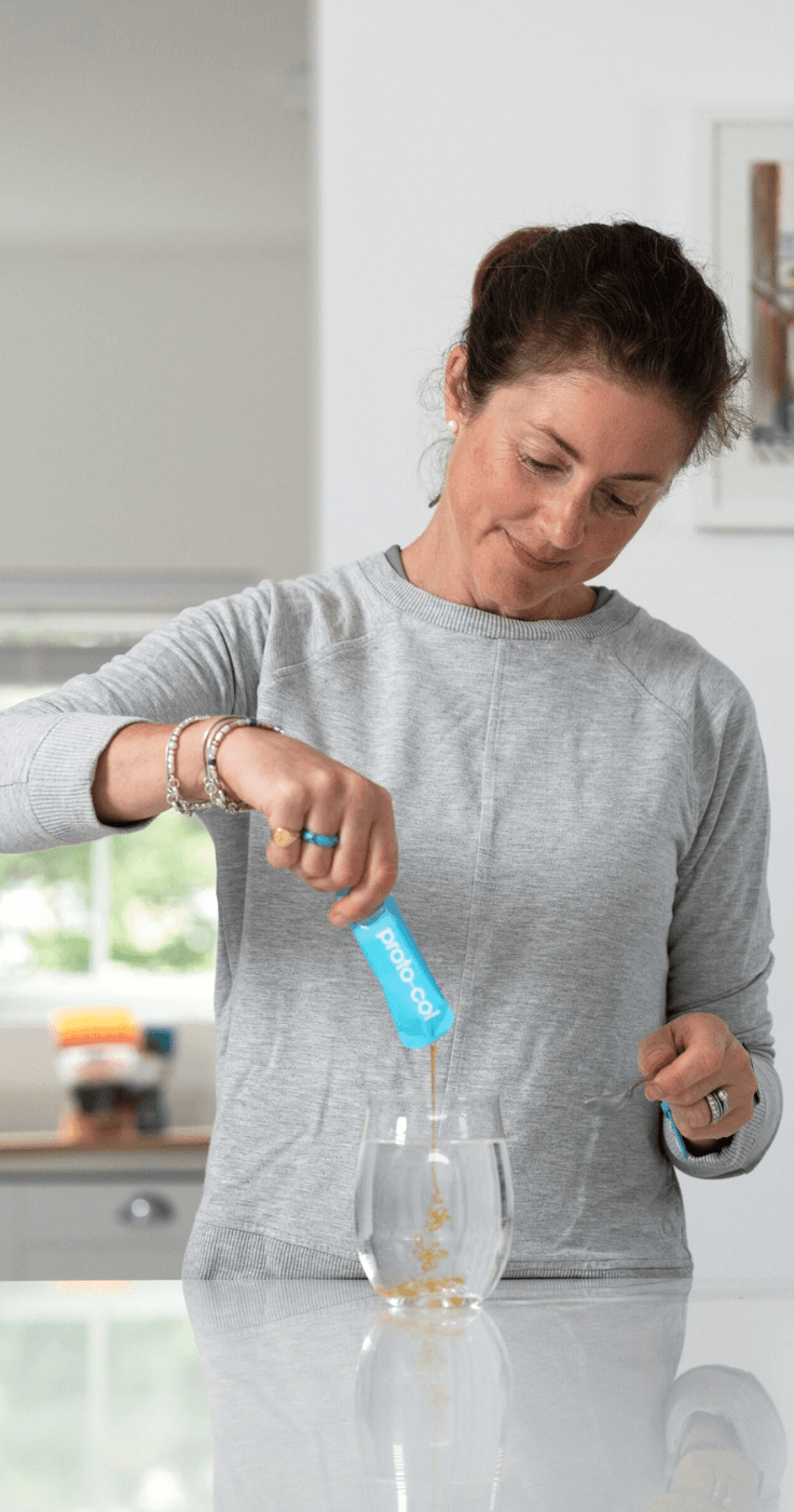 A woman in a grey sweatshirt preparing a glass of collagen drink by pouring a sachet of Proto-col marine collagen into water. She is smiling and stirring the mixture, representing a daily wellness routine. Keywords: Collagen Drink UK, Beauty Supplements for Skin, Health and Wellbeing.