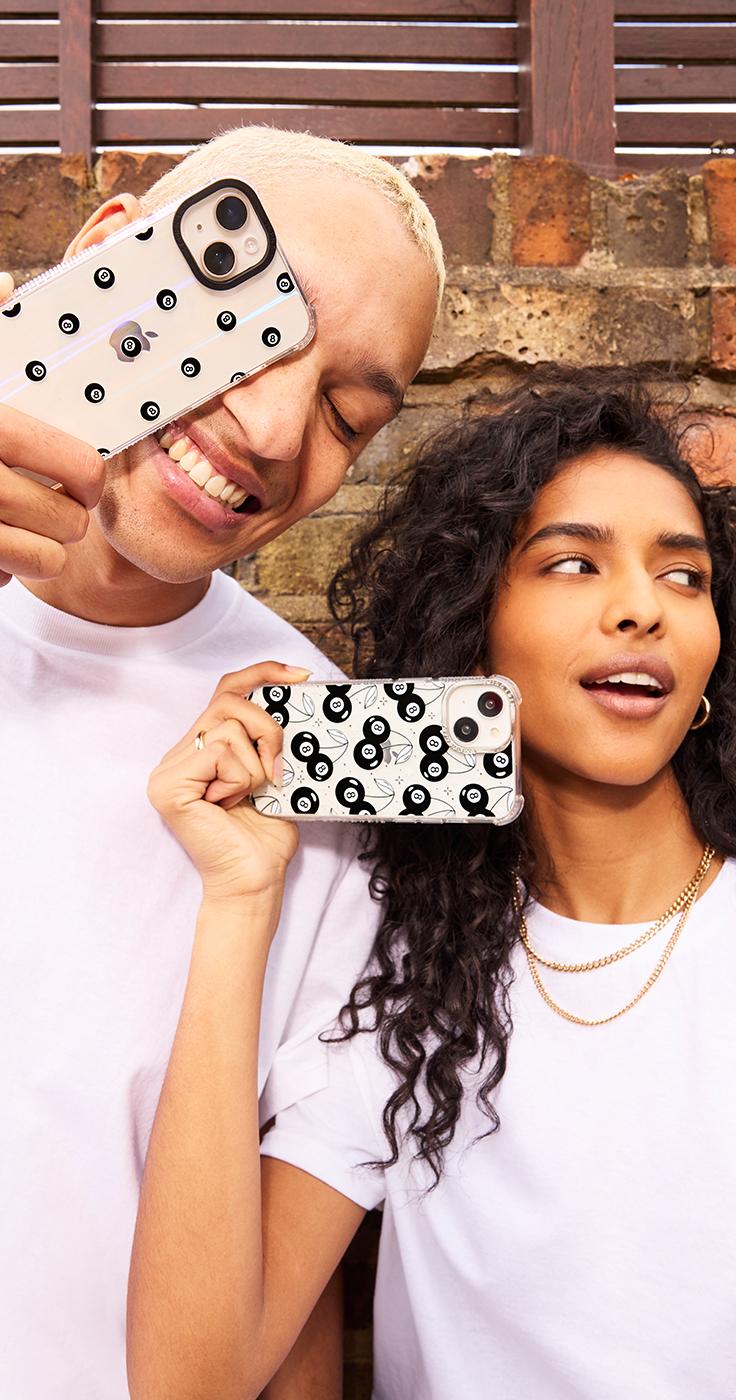 Two people smiling and posing with phones in cases featuring a black and white 8-ball pattern. They are wearing white shirts and standing against a brick wall.