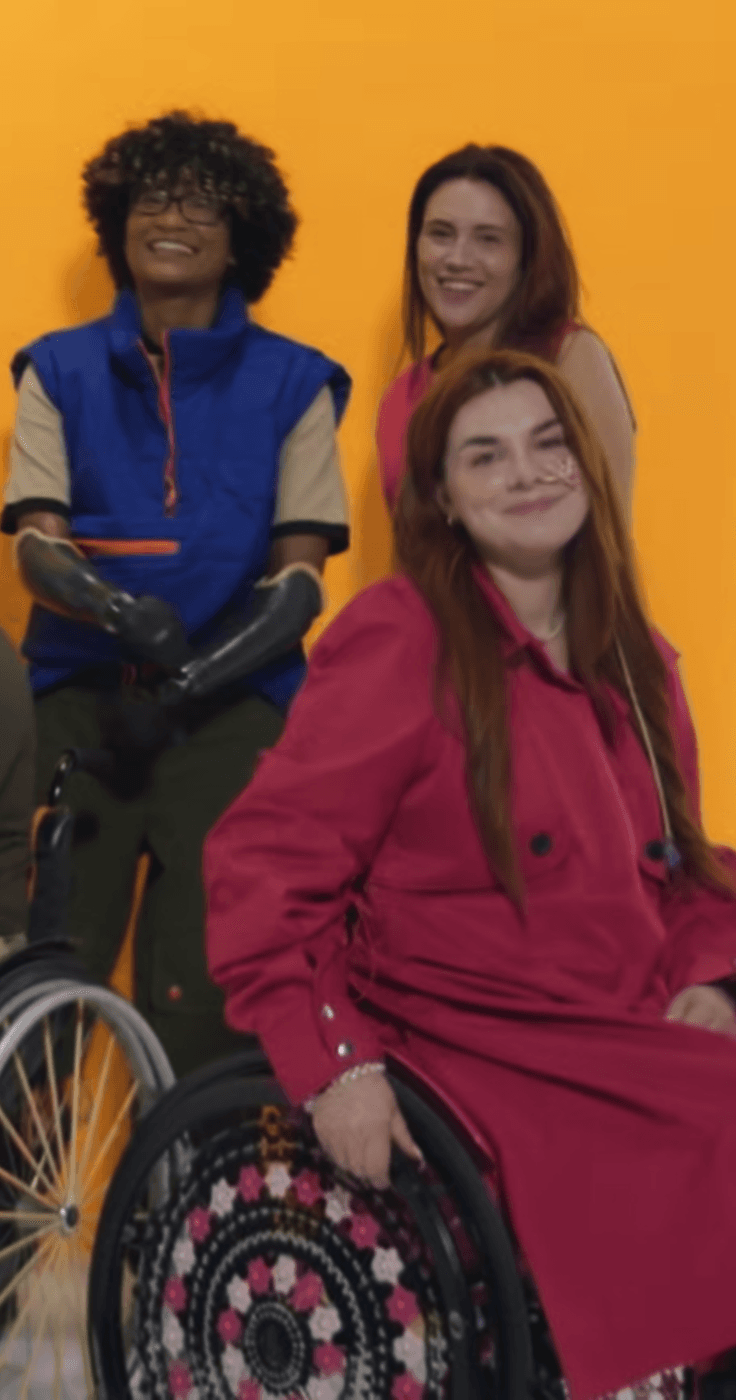  Three people are smiling in front of an orange background. One person in a wheelchair wears a red coat, another with curly hair and prosthetic arms wears a blue vest, and the third person with long brown hair stands behind them.