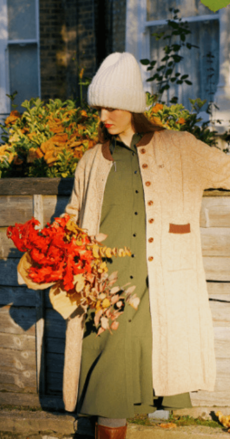 A woman in a white knit hat and warm autumnal coat holds a bouquet of bright red leaves, embodying a comfortable and fashionable countryside style ideal for Purpl members.