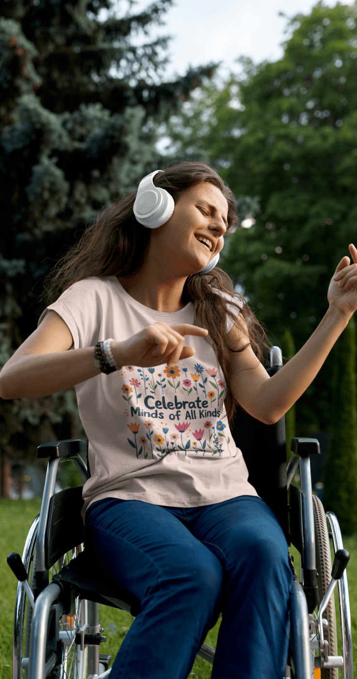 A woman in a wheelchair enjoys music in a park, wearing headphones and dancing. She has long brown hair and a t-shirt that reads "Celebrate Minds of All Kinds" with floral designs. Trees and greenery are in the background.