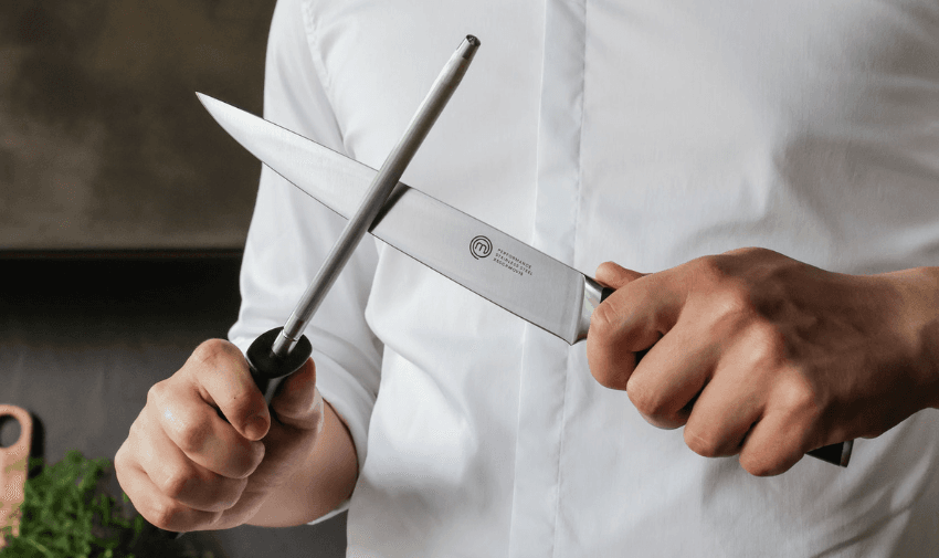 A chef in a white uniform sharpens a professional MasterChef knife using a honing steel, demonstrating precision and quality in culinary tools. Save on Kitchen Essentials for Disabled People.