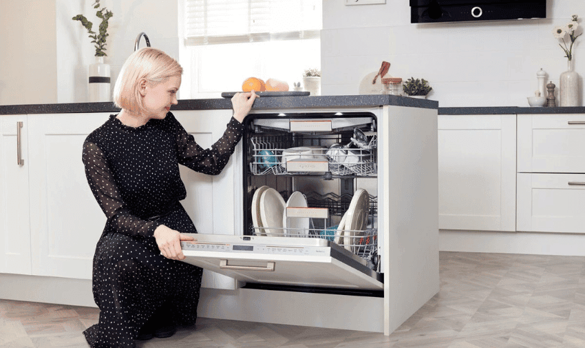  A woman with short blonde hair, wearing a black dress with white polka dots, is kneeling and opening a dishwasher in a modern kitchen. The dishwasher is partially filled with plates and other dishes. The kitchen has white cabinets and a light gray floor.