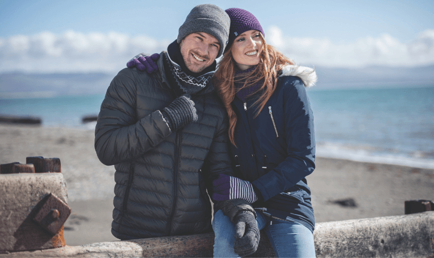 A couple dressed in winter clothing, including hats and gloves, stand close together on a beach. The man leans on a wooden post, and the woman has her arm around him. The sea and cloudy sky are in the background.