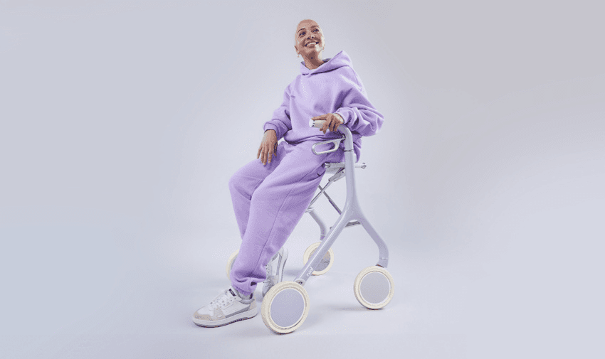 A person smiles while sitting on a stylish lavender Zeal walking aid. They wear a matching lavender tracksuit and white sneakers, with a neutral background highlighting the modern design of the mobility device.