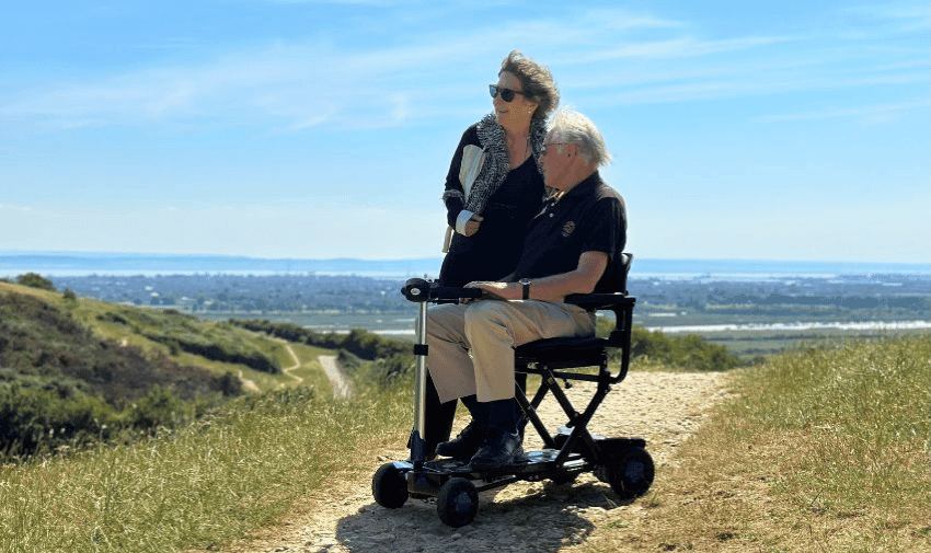  A woman stands next to an older man seated on a mobility scooter on a dirt path atop a hill. Both are dressed in casual clothes and sunglasses. The horizon shows a clear sky and a blur of distant landscape beneath them. They appear to be enjoying the scenery.