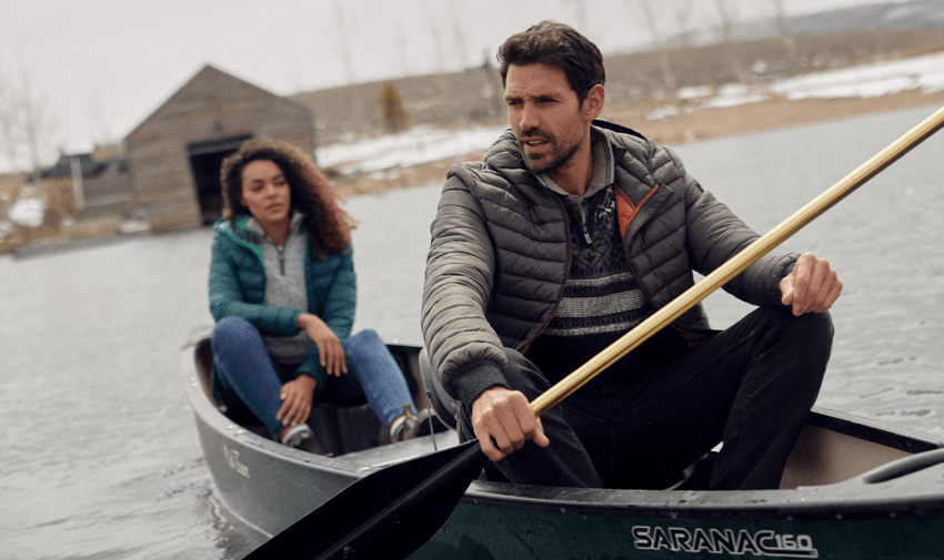 A man and a woman are canoeing on a calm lake. The man is paddling while the woman sits behind him. Both are wearing jackets, and there is a wooden cabin and snowy landscape in the background.