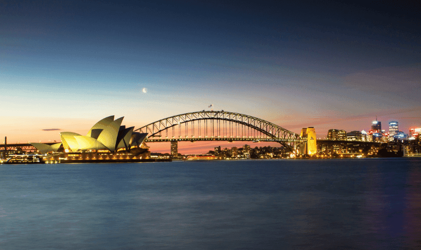 A panoramic view of Sydney Harbour featuring the illuminated Sydney Opera House on the left, the Harbour Bridge in the center, and a cityscape with buildings on the right, all set against a twilight sky with a crescent moon.