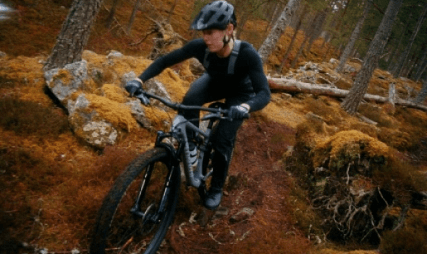 A person wearing a helmet and dark clothing rides a mountain bike on a narrow, rugged trail through a dense forest with moss-covered ground and rocks. The scene suggests action and focus amidst natural surroundings.