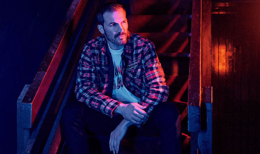 A man sitting on stairs in moody lighting, wearing a plaid jacket and graphic T-shirt from Mainline Menswear. Showcasing Purpl's disabled discounts for trendy and high-quality men's clothing in the UK.