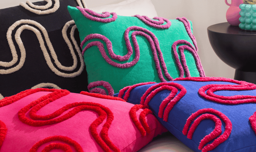 A collection of colourful throw pillows featuring a bold wavy pattern in contrasting colours. The pillows are placed on a bed, with one in teal, one in black, one in pink, and one in blue. A black round side table with decorative items is visible in the background.