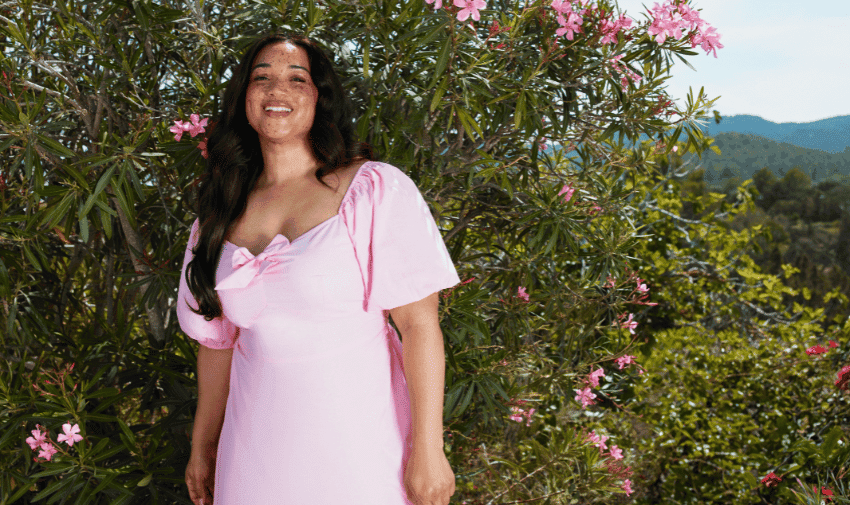 A woman in a light pink dress stands smiling in front of green shrubs with pink flowers. The background shows a scenic view of mountains and a clear blue sky.