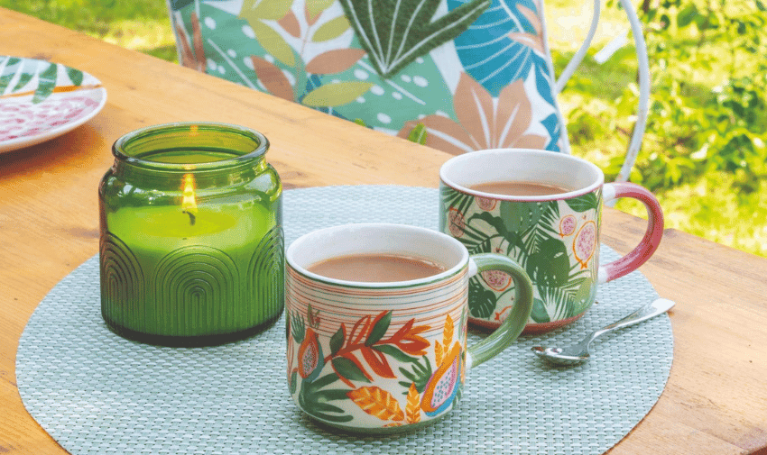 A cosy outdoor table setting featuring two colorful mugs filled with tea or coffee, placed on a light blue woven placemat. A lit green candle in a glass jar and a spoon rest nearby. The background shows a brightly patterned cushion and leafy greenery.