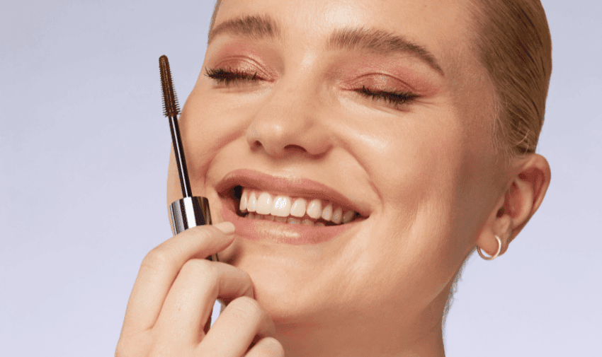 A person with light skin and blonde hair smiles with eyes closed while holding a mascara wand close to their face. They are wearing pink eyeshadow and small hoop earrings. The background is a light shade of blue.