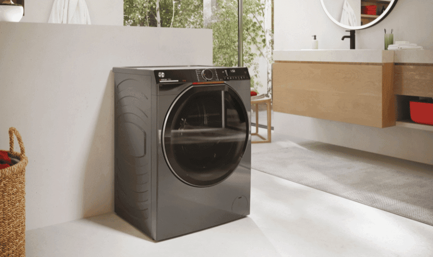 A Hoover washing machine with a modern, front-loading design and a sleek, dark gray exterior is situated in a clean, minimalistic laundry space. A wicker basket is partially visible to the left, and a contemporary bathroom vanity with a round mirror and sink can be seen in the background.