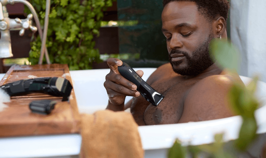 A man is relaxing in a bathtub while using a Manscaped electric grooming tool to shave his chest.