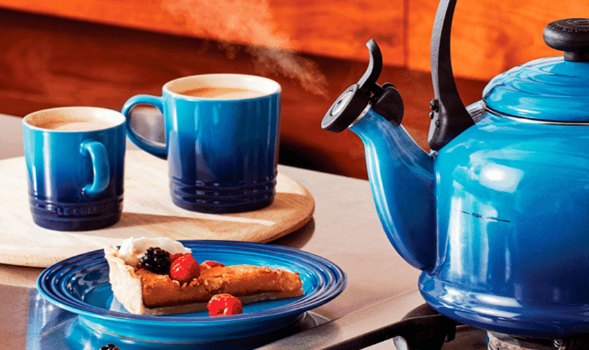 A Le Creuset vibrant blue kettle on a stovetop with steam rising from its spout. On the counter, a matching blue plate holds a slice of toast topped with berries. Two blue mugs filled with frothy coffee sit on a wooden board in the background.