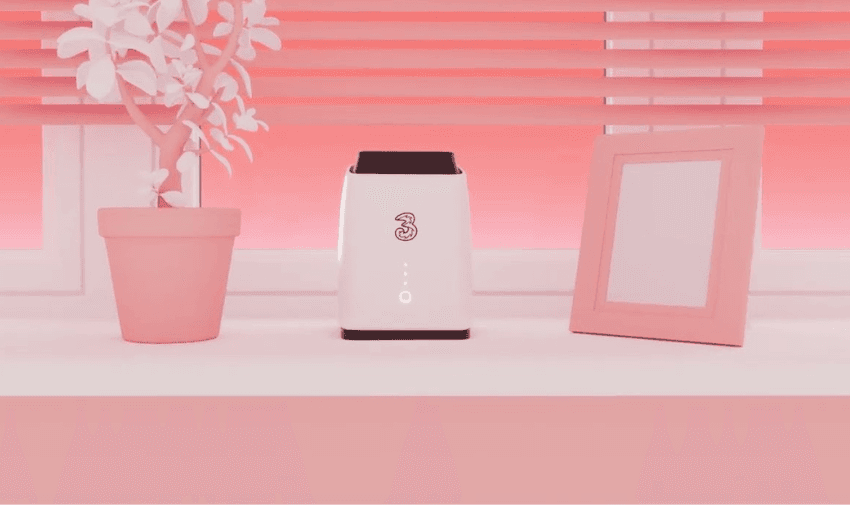 A sleek white 5G broadband hub with the Three logo on a windowsill, surrounded by a pink potted plant and a photo frame.