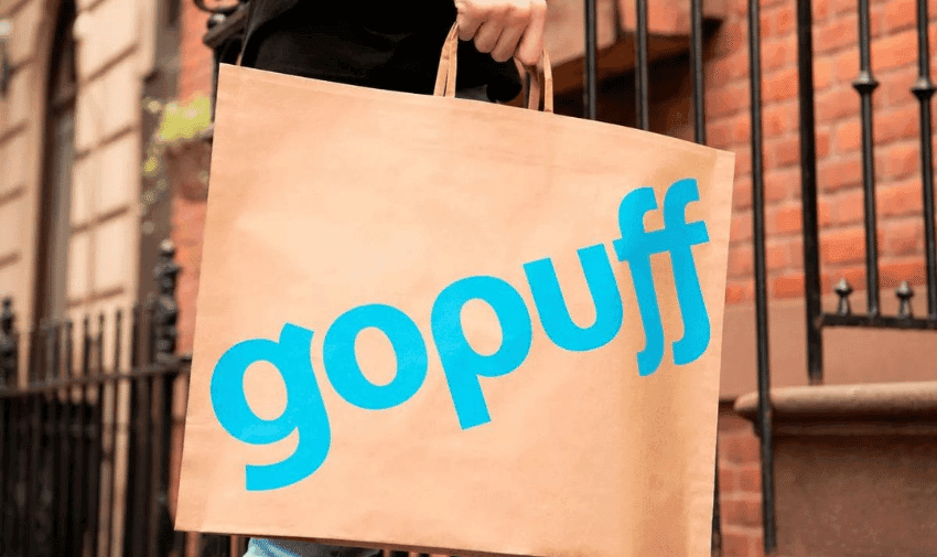 A person is carrying a brown paper bag with the blue "GoPuff" logo. The background shows a brick building and a metal railing, indicating an urban residential setting.