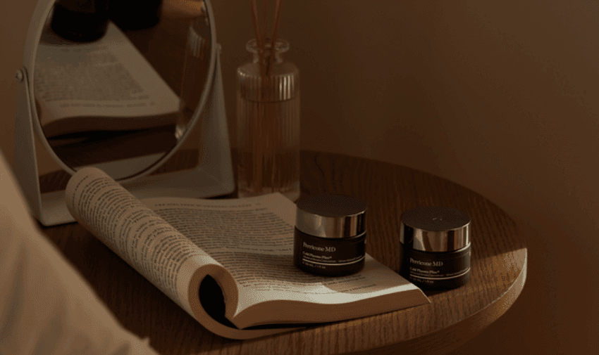 A wooden table with an open book, two jars of Perricone MD skincare products, and a small round mirror. In the background, there's a reed diffuser with amber liquid. Warm, soft lighting creates a cozy, serene atmosphere.