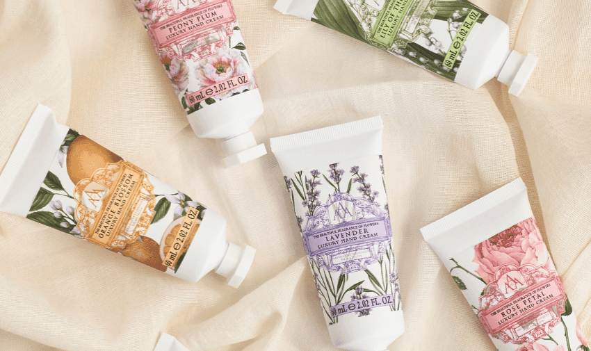 Five decorative tubes of Somerset Toiletry Co hand cream are displayed on a soft beige fabric. The tubes are labeled with various floral designs and scents, such as Lavender and Rose. Each tube has a different pastel colour theme with floral illustrations.