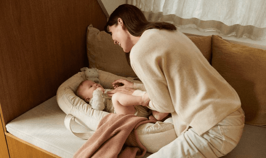 A mother smiling while gently playing with her baby lying in a cosy nest on a bench. Features Baby Care Discounts and Disabled Family Discounts.