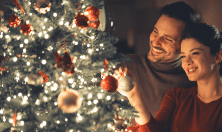 A couple decorates a sparkling Christmas tree together, surrounded by glowing fairy lights and festive ornaments, showcasing holiday joy.