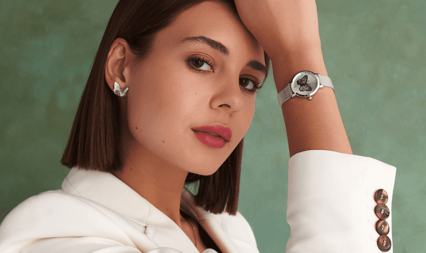 A woman in a white jacket poses with her hand resting on her head. She wears a watch with a portrait on the dial, butterfly-shaped earrings, and her hair is straight and shoulder-length. The background is a textured green.