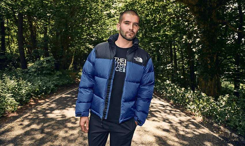  A person wearing a blue puffer jacket and a black shirt stands on a path surrounded by dense green trees in a forest. The sunlight filters through the leaves, casting dappled shadows on the ground.