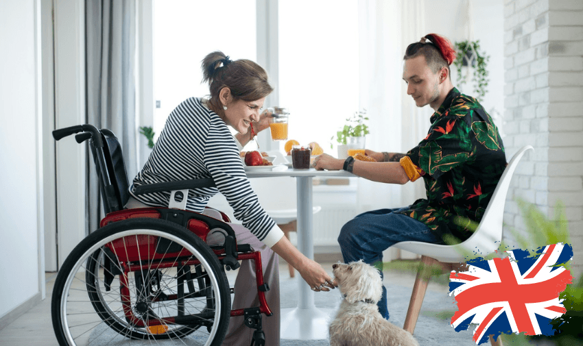 A woman in a wheelchair and a man are sitting at a dining table, enjoying a meal together. The woman, who looks happy, is wearing a striped shirt and petting a small dog. The man, dressed in a colourful shirt, appears to be mid-conversation. The setting is a bright, modern room reflecting inclusivity and warmth.