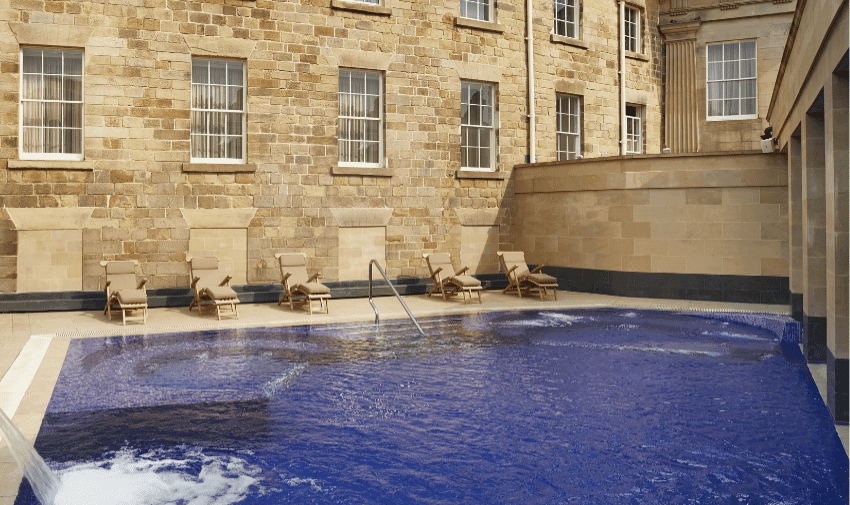 An outdoor pool beside a historic stone building. Five lounge chairs with cushions are set up along the pool's edge. The building has multiple large windows and the pool water appears calm with slight ripples.