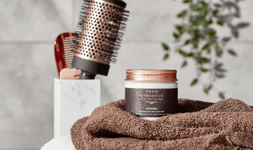 A jar of Grow Gorgeous Intense hair mask sits on a brown towel. Next to it are a cylindrical copper hairbrush with one black and one brown handle protruding from a white marble holder. In the background, a leafy plant is partially visible.