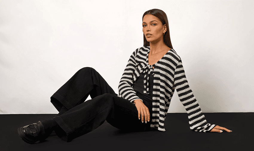 A person with long, brown hair sits on the floor against a plain white backdrop. They wear a black and white striped sweater, black pants, and black shoes. The person's legs are extended, with one leg bent and the other outstretched. The person looks relaxed.