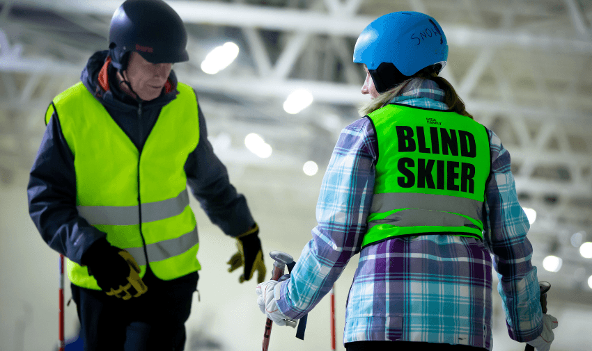 A person wearing a blue helmet and a vest labeled "Blind Skier" holds ski poles while talking to another person in a black helmet and yellow vest. They are standing indoors on a snowy surface.