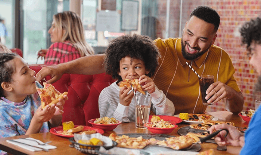 A family enjoys a meal at a Pizza Hut restaurant, smiling and sharing pizza. A father in a mustard shirt playfully feeds his child while another child takes a bite of pizza. Plates of salad, corn, and drinks are on the table. Pizza Hut UK restaurant savings, family dining deals, disabled discount offers.