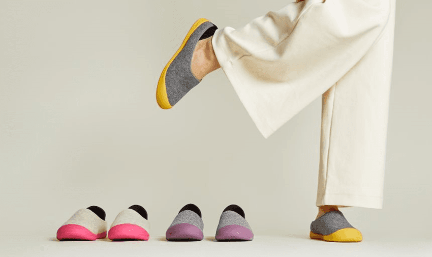 a person is wearing a pair of grey mahabis slippers with yellow soles and is holding on leg up - there are two other pairs on the floor with pink and purple soles.