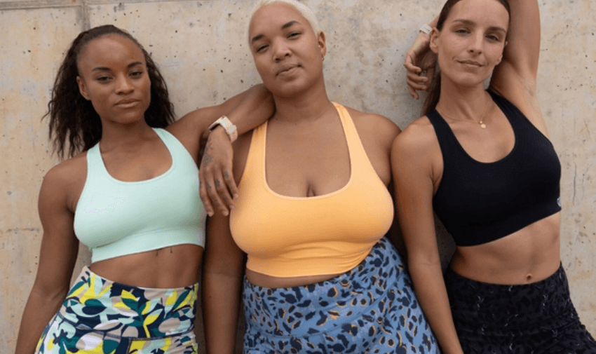 Three women wearing athletic wear pose confidently against a concrete wall. They are dressed in colourful sports bras and patterned leggings, showcasing a diverse range of body types and expressions of empowerment.