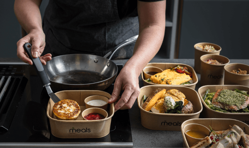 A person plating a golden-brown dish from a pan into a meal container, surrounded by pre-prepared dishes in eco-friendly packaging with the "Meals" logo.